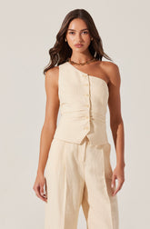 A woman is wearing a sleeveless, off-white, beige-colored vest with front buttons and matching high-waisted, wide-leg pants. The outfit appears to be made of a lightweight, textured fabric. She has accessorized with gold hoop earrings and a chunky gold chain necklace.